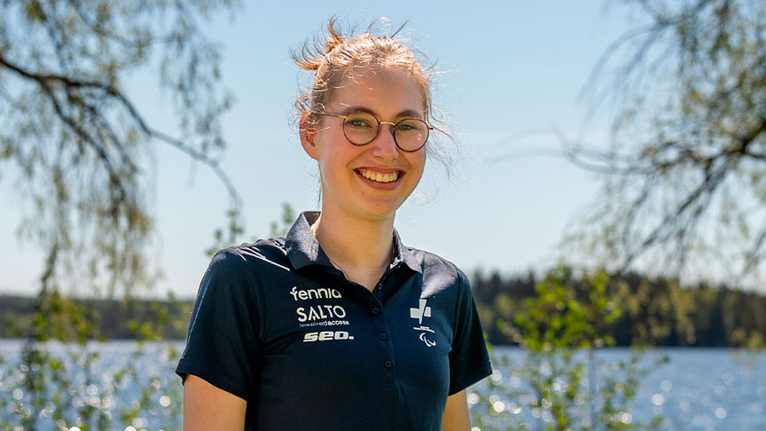 Aamu Hulmi hymyilemässä kameralle ulkona aurinkoisessa säässä. Taustalla vihreää kasvustoa ja vesistö.